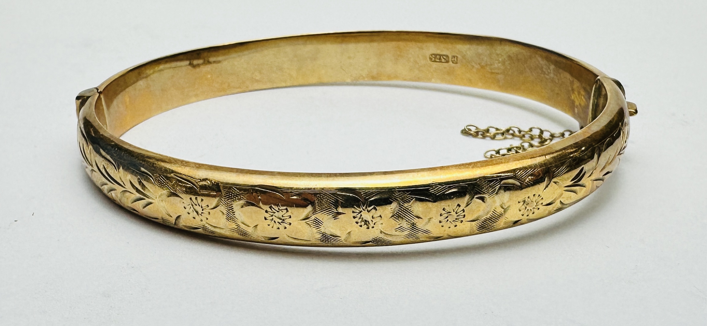 A 9CT GOLD HINGED BANGLE WITH SAFETY CHAIN AND CHASED DECORATION.