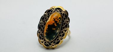 A YELLOW METAL (WORN MARKS) OVAL AMBER TYPE SET RING IN FILIGREE MOUNT - SIZE L/M.