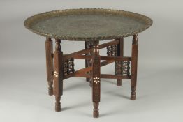 AN ISLAMIC ENGRAVED BRASS TRAY TABLE, with folding bone inlaid wooden legs, tray 66cm diameter.
