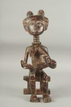 A CARVED AFRICAN FIGURE OF A WOMAN SITTING ON A BENCH, feeding a baby. 14ins high.