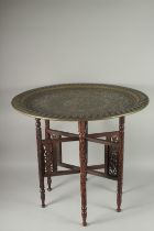 A LARGE ISLAMIC EMBOSSED AND CHASED BRASS TRAY TABLE, with folding carved wood legs, tray 76cm