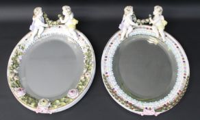 Two oval floral encrusted porcelain mirrors surmounted with cherubs holding garlands, bearing