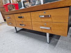 A LOW SIDE CABINET WITH THE RECTANGULAR TEAK TOP OVER SIX DRAWERS WITH CHROME HANDLES, THE SIDES