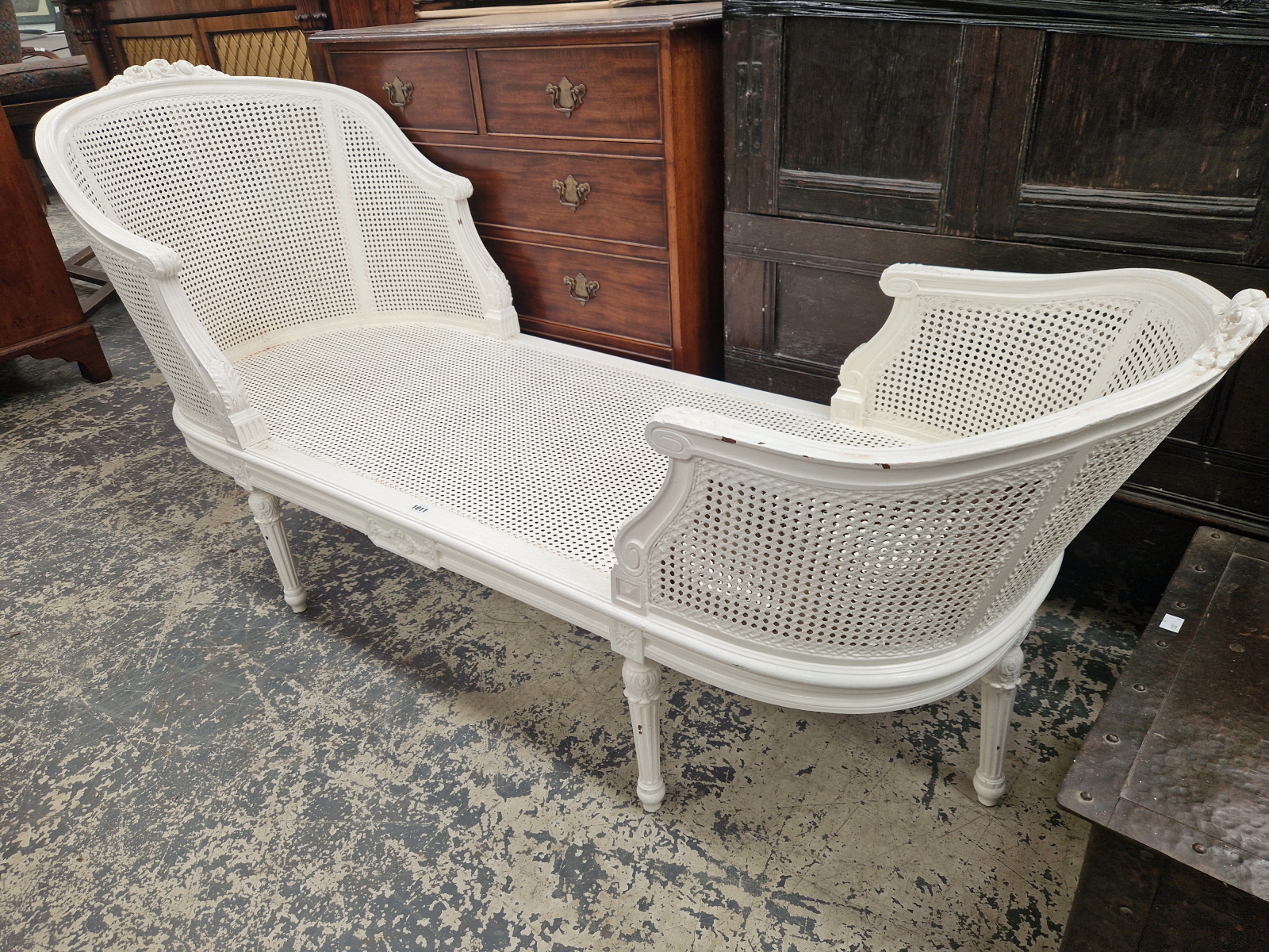 A WHITE PAINTED CANED CHAISE LONGUE, THE ROUNDED ENDS CRESTED BY CARVED FLOWERS, THE FLUTED