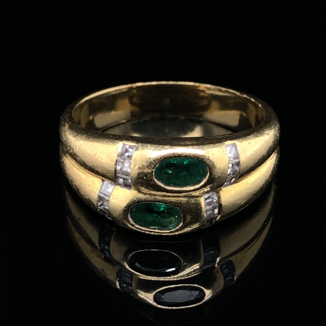 AN EMERALD AND DIAMOND RING. THE TWO OVAL CUT EMERALDS FLANKED WITH A CHANNEL SET OF PRINCESS CUT