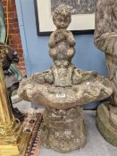 A WEATHERED COMPOSITE STONE FOUNTAIN, THE SHELL BOWL BACKED BY A CHILD SATYR PLAYING PAN PIPES.