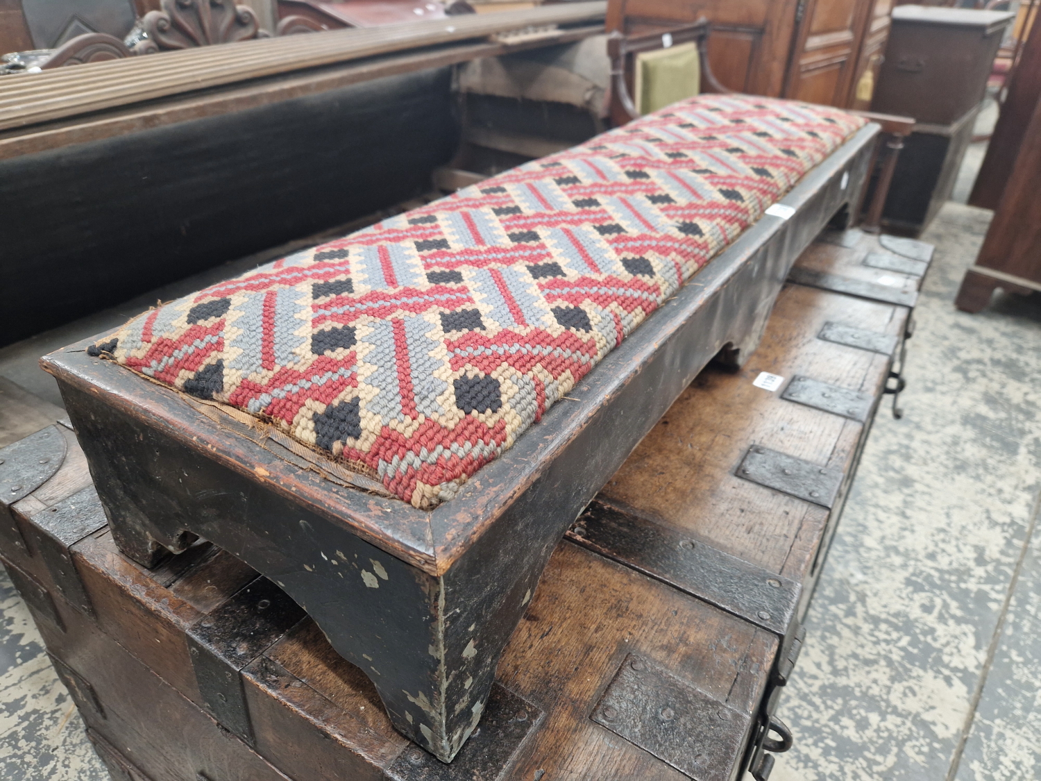 A BLACK PAINTED PINE AND TOLE FOOT REST WITH A NEEDLE WORKED DROP IN TOP.   W 109cms.