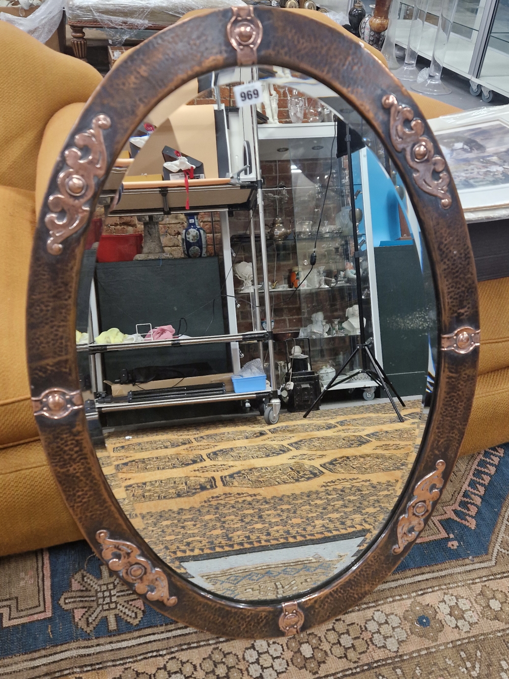 A BEVELLED GLASS OVAL MIRROR IN A HAMMERED COPPER FRAME APPLIED WITH ALTERNATING CABOCHON AND SCROLL