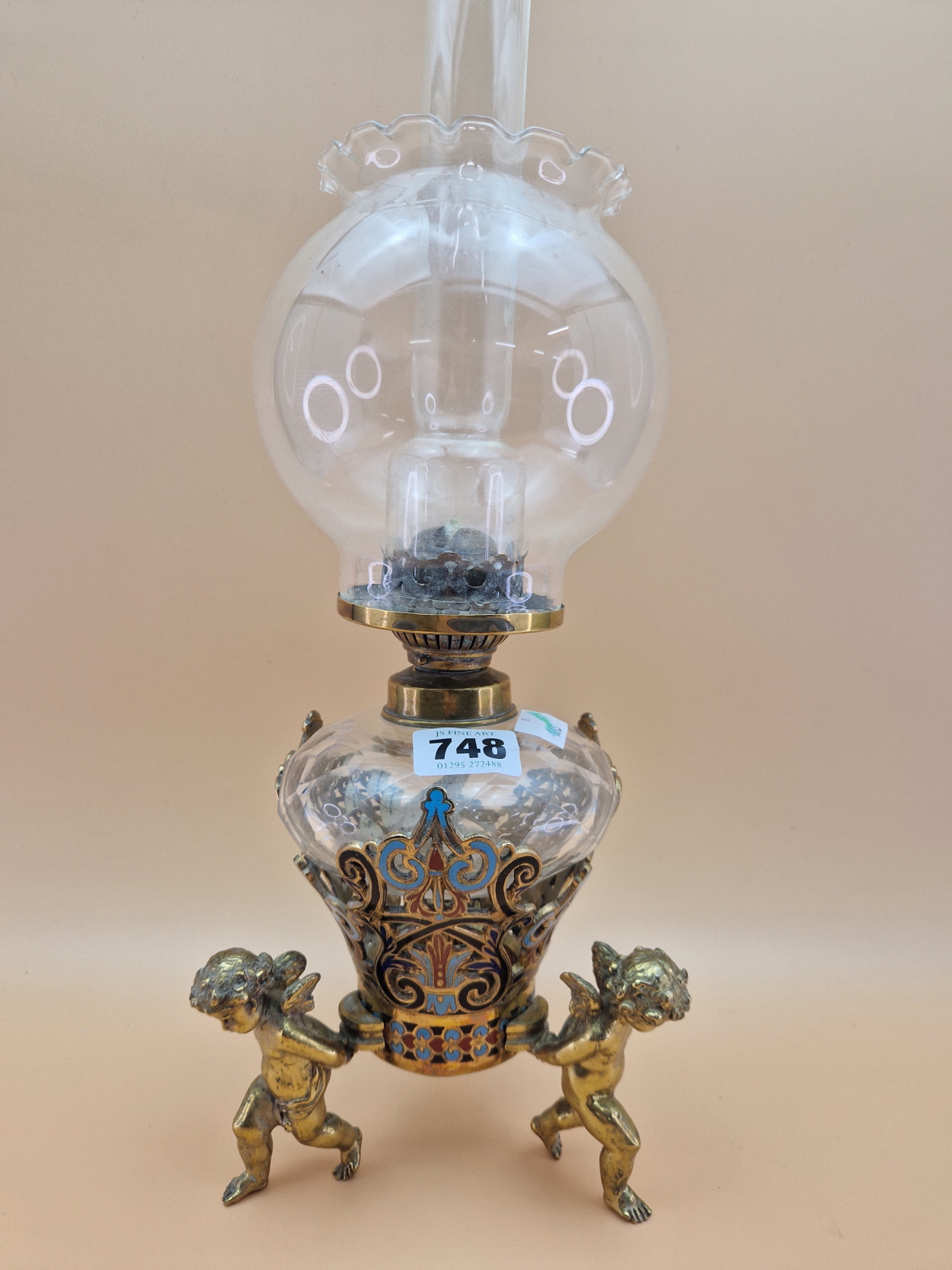 A LATE 19th C. FRENCH CLEAR CUT GLASS AND CHAMPLEVE ENAMEL OIL LAMP SUPPORTED BY THREE BRASS CUPIDS