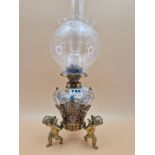 A LATE 19th C. FRENCH CLEAR CUT GLASS AND CHAMPLEVE ENAMEL OIL LAMP SUPPORTED BY THREE BRASS CUPIDS