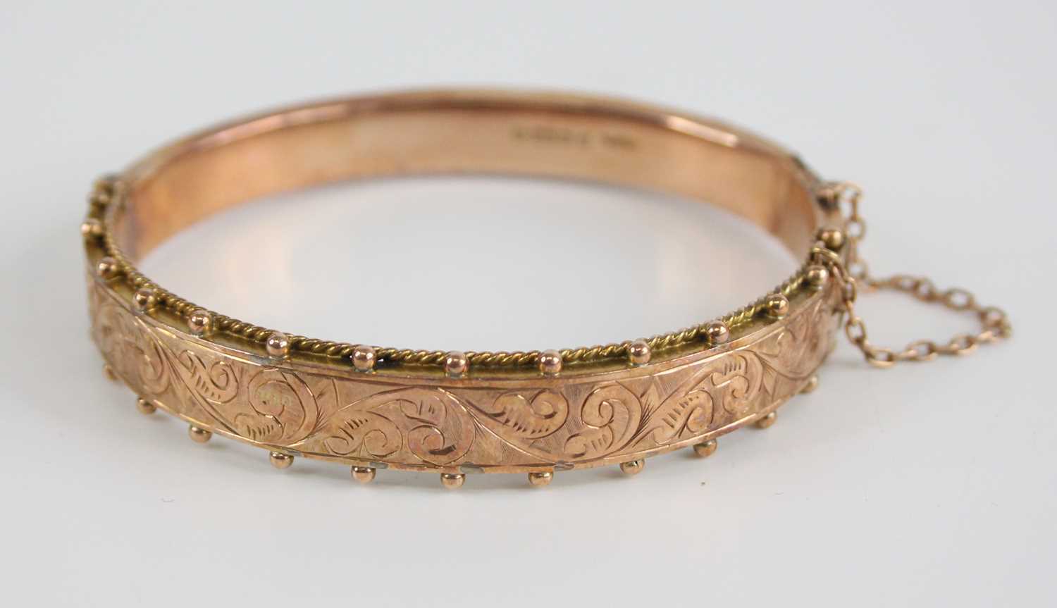 An Edwardian 9ct gold hinged hollow bangle, one side having engraved scroll leaf decoration within a