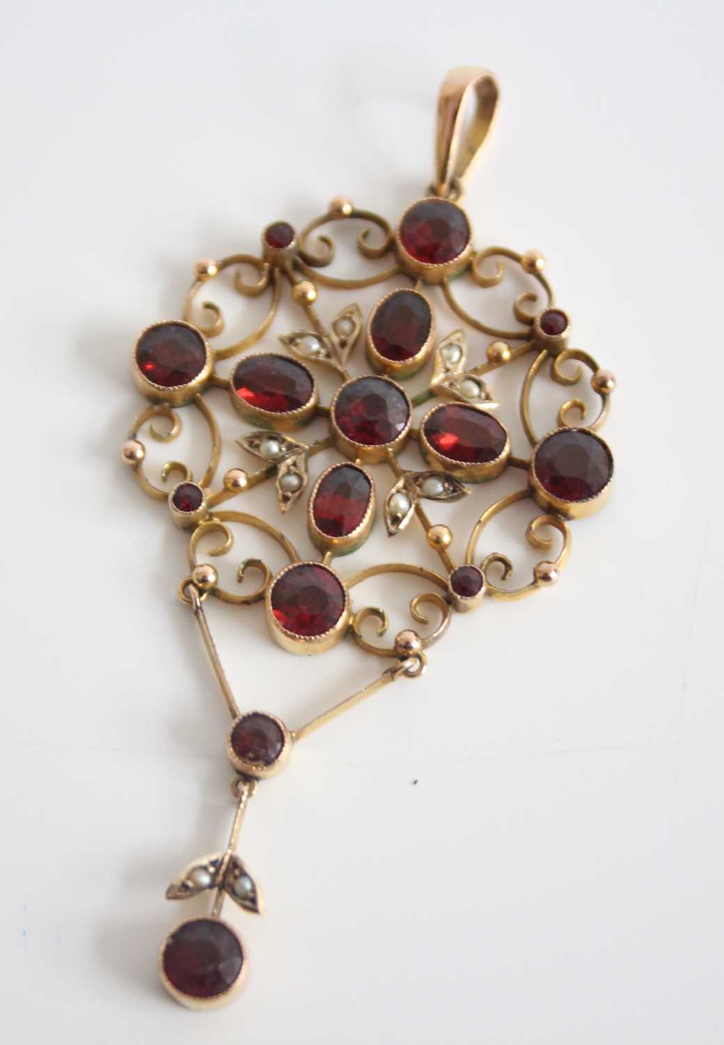 A yellow metal Edwardian openwork garnet and seed pearl pendant, featuring ten round and four oval