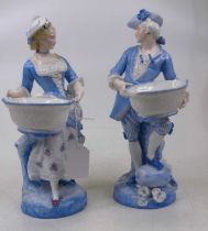 A pair of porcelain figures of a gentleman and lady, each shown standing holding a basket, height