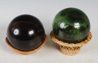 Two glass floats set on wicker baskets, one green the other brown, approximately 11.5cm diameter.