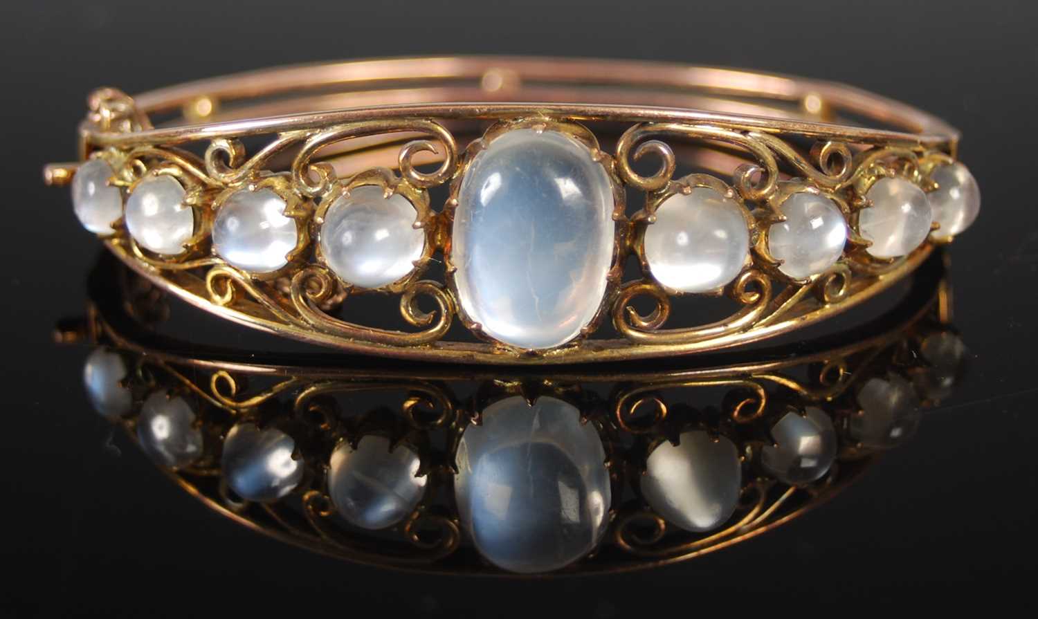 A yellow metal and moonstone hinged bangle, centred an oval cabochon cut moonstone flanked on either