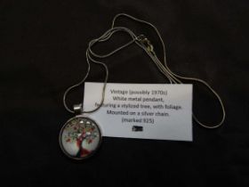 A white metal pendant containing artwork of stylised tree mounted on white metal chain