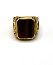 A yellow metal and tigers eye gents ring, set with a smooth tigers eye plaque, the shoulders with