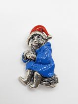 A silver and enamel Paddington Bear brooch, wearing a red hat and blue raincoat, eating a