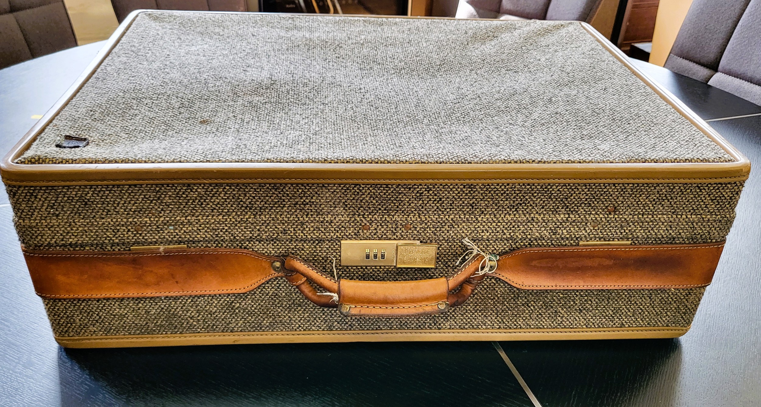 A Hertman suitcase, covered in grey fabric with brown leather straps.