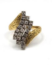A yellow metal and diamond crossover ring, the textured shoulders set with three rows of round