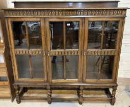 A 1920's Jacobean style oak display cabinet enclosed by 3 glazed doors, on short barley twist legs