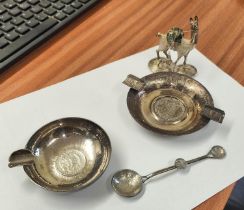 A two white metal South American coins set into ashtrays, a similar spoon and similar camel weight