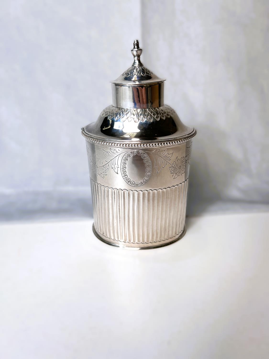 An oval white metal tea caddy with chased decoration, beaded rim and flared lower section marked for