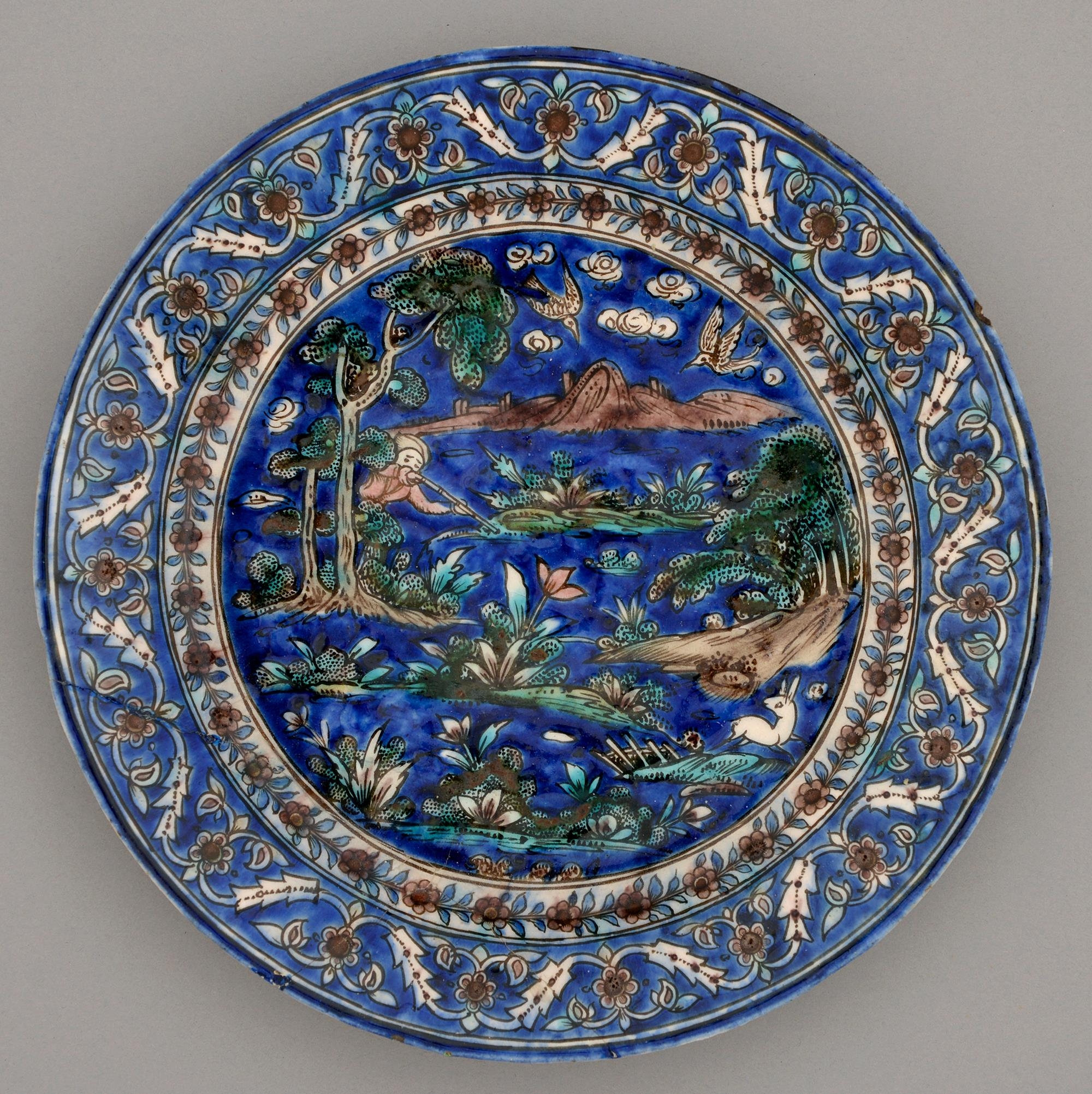 A Persian frit paste dish, Qajar dynasty, 19th c, painted with a hunter, deer and birds amidst