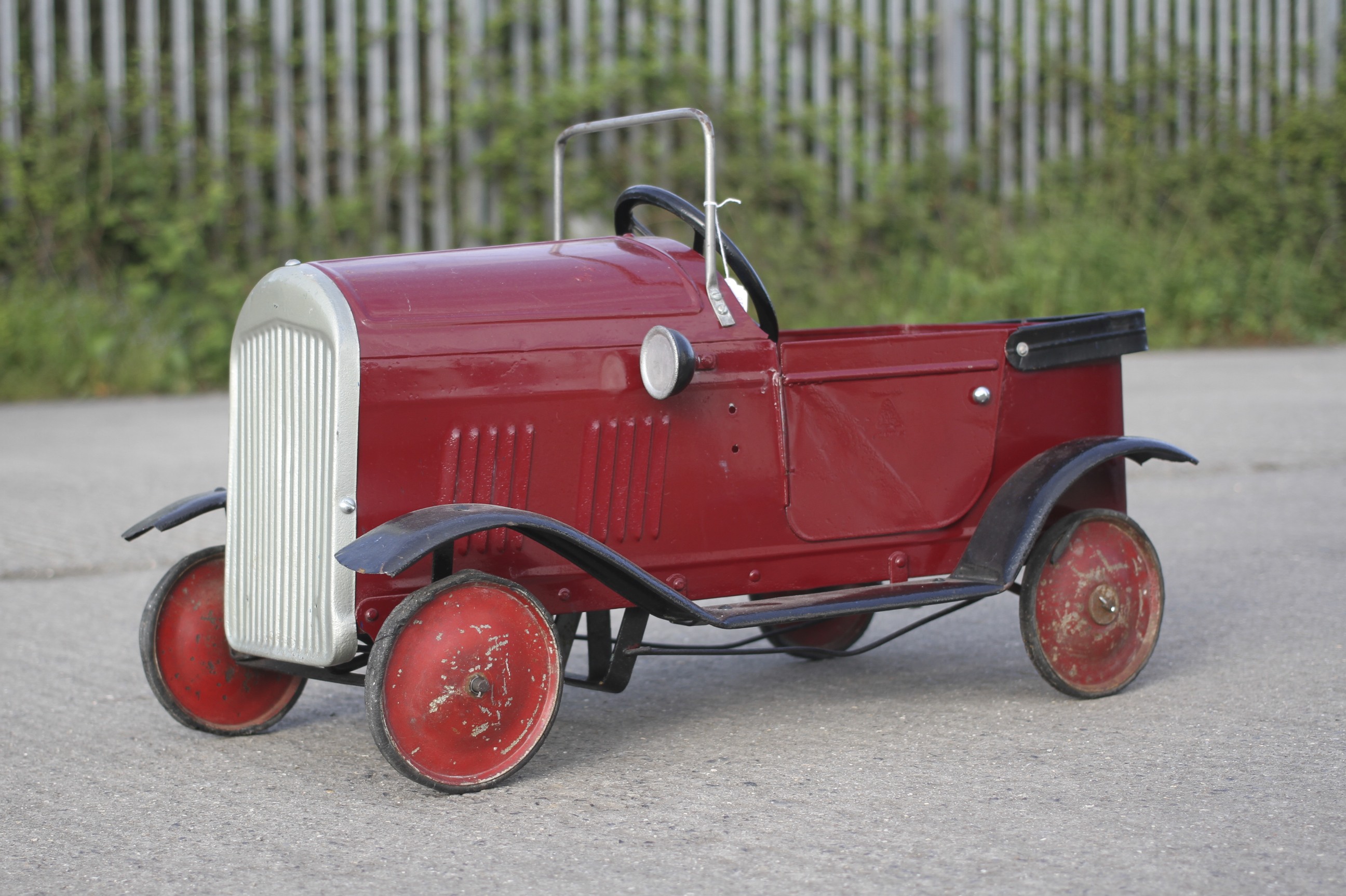 A circa 1930s tin plate pedal car. Red body with black trim and red wheels, H49cm x L77cm.