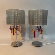 A pair of white metal table lamps hung with pendant prismatic drops in clear and coloured glass,