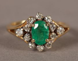 AN EMERALD AND DIAMOND CLUSTER RING, the oval faceted emerald claw set, raised against a surround of