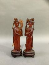 A pair of Chinese carved amber effect figures of women in robes holding flowers, on carved stands,