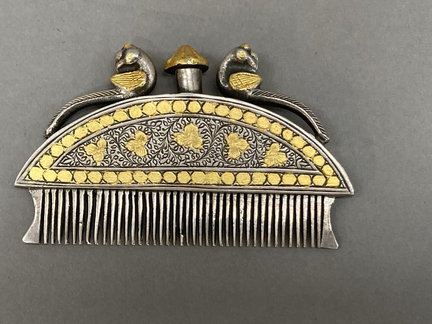 An Indian, white metal moustache comb with gilt metal inlay, the finial, surmounted by two peacocks,