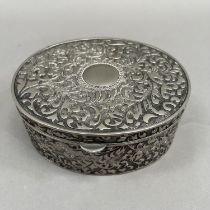 An oval box in white base metal with hinged lid, all over foliate chasing and an open circular