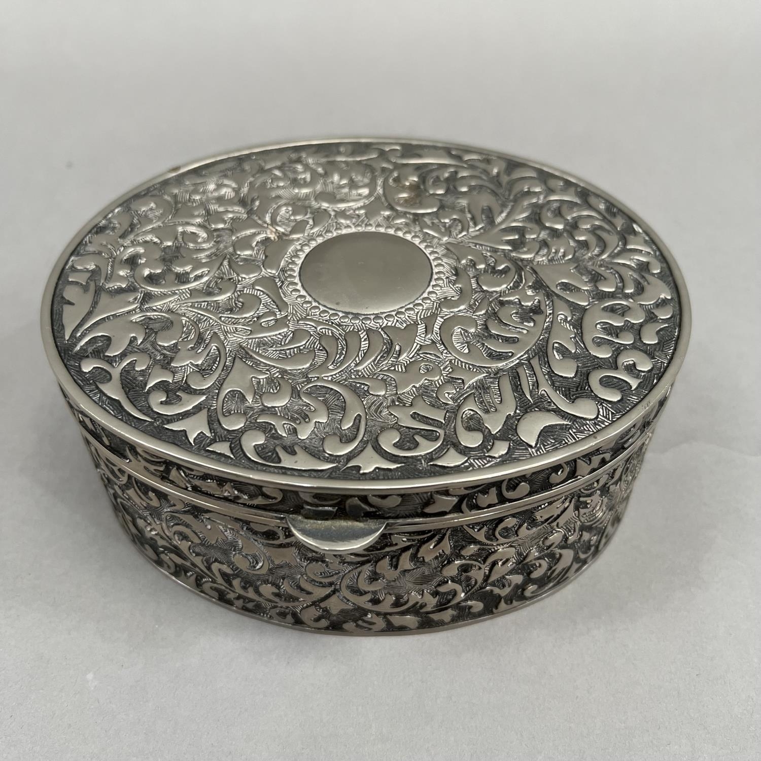 An oval box in white base metal with hinged lid, all over foliate chasing and an open circular