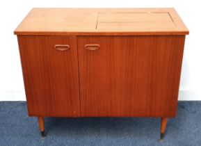 TEAK SEWING MACHINE CABINET with a fold over flap top above a pair of cupboard doors, standing on