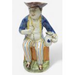 A Prattware Pottery ‘Sailor’ toby jug, c1790-1810, seated holding a jug, striped britches, blue coat