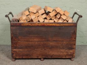 A panelled log basket with metal surround, containing a large quantity of pre-cut logs. Bearing