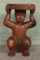 A carved hardwood plant stand depicting a tribal figure holding a board. Height: 48cm. Missing
