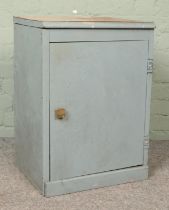 A grey painted wooden work cabinet, with hinged door revealing a set of shallow drawers. Contains