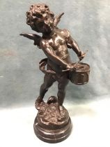 A bronze figure of Cupid playing a drum standing on a naturalistic base, with dark patina on a