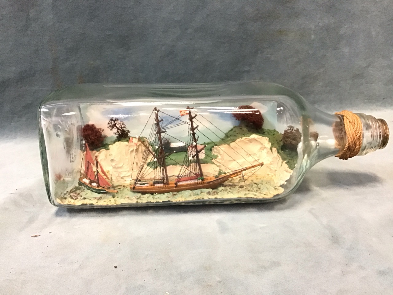 A ship in a bottle diorama with two sailing boats below a clifftop village in a square bottle - - Image 3 of 3