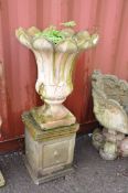 A WEATHERED COMPOSITE GARDEN URN ON STAND with a flower head shape and Acanthus leaf detailing on