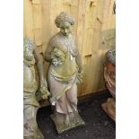 A WEATHERED COMPOSITE GARDEN FIGURE in the form of a Roman lady resting by a tree stump holding