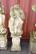 A WEATHERED COMPOSITE GARDEN FIGURE of embracing lovers standing on a ornately decorated base
