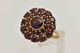 A GARNET CLUSTER RING, the central circular cut garnet in a collet setting within a two tier claw