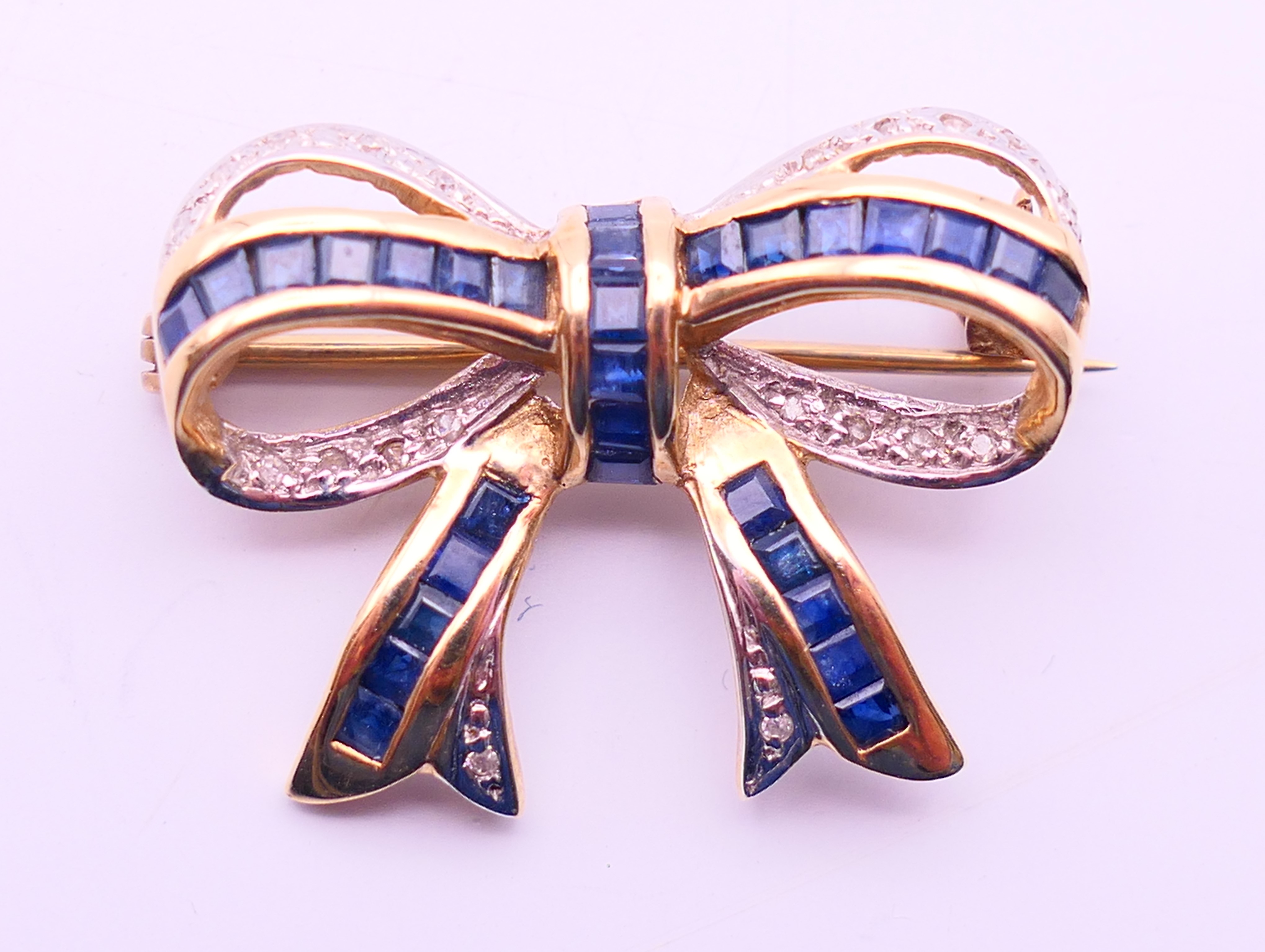 A 9 ct gold bow brooch set with mid-blue baguette sapphires and diamonds. 2.5 cm x 2 cm. 3.