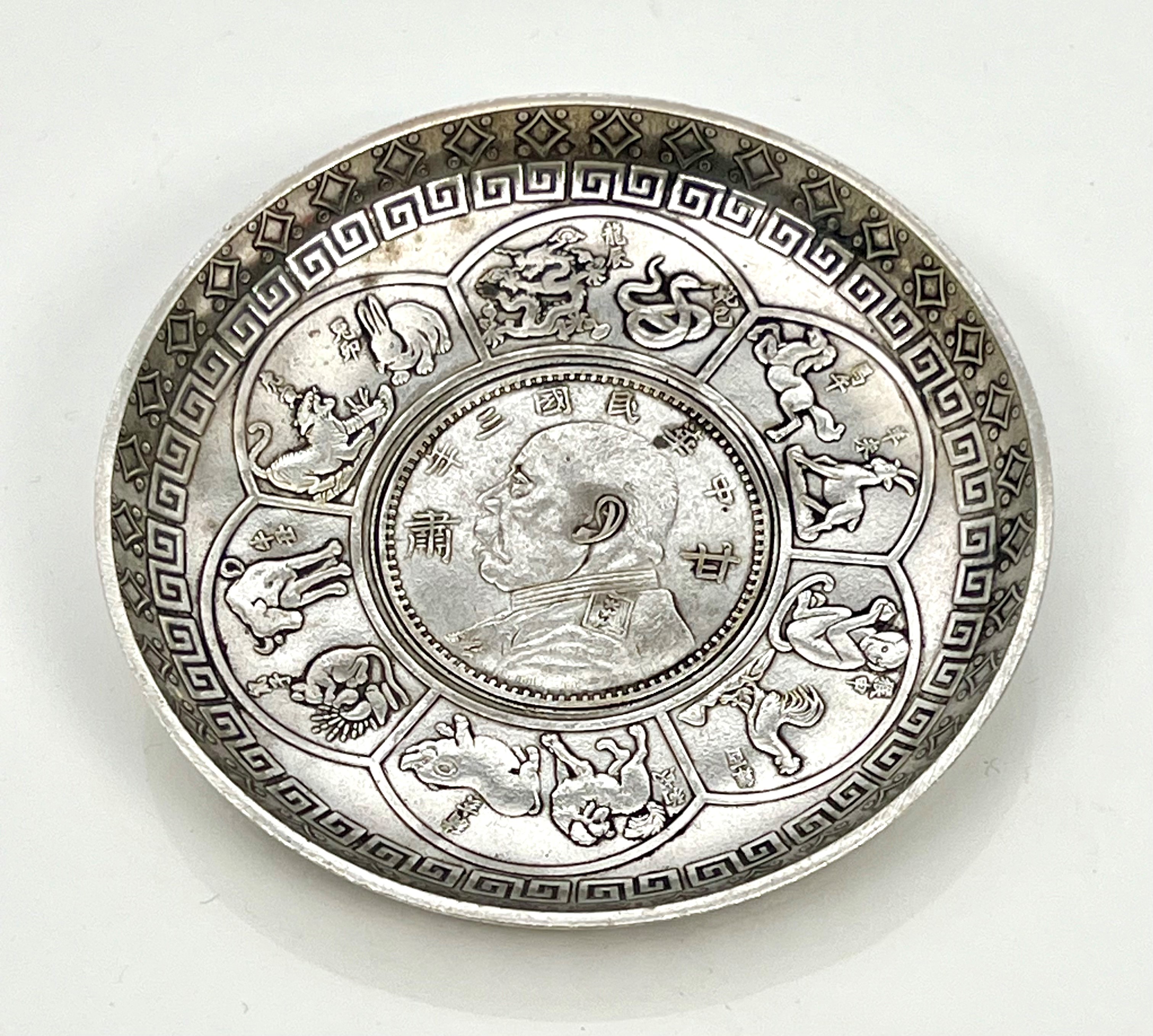 A Chinese white metal zodiac dish - inset with a Chinese $1 one dollar coin, within a flowerhead