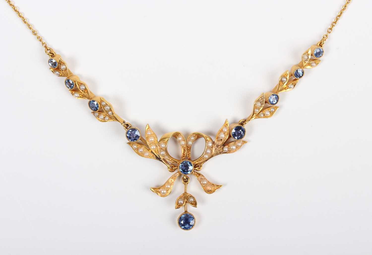 An Edwardian gold, sapphire and seed pearl necklace, the front designed as a tied bow with foliate