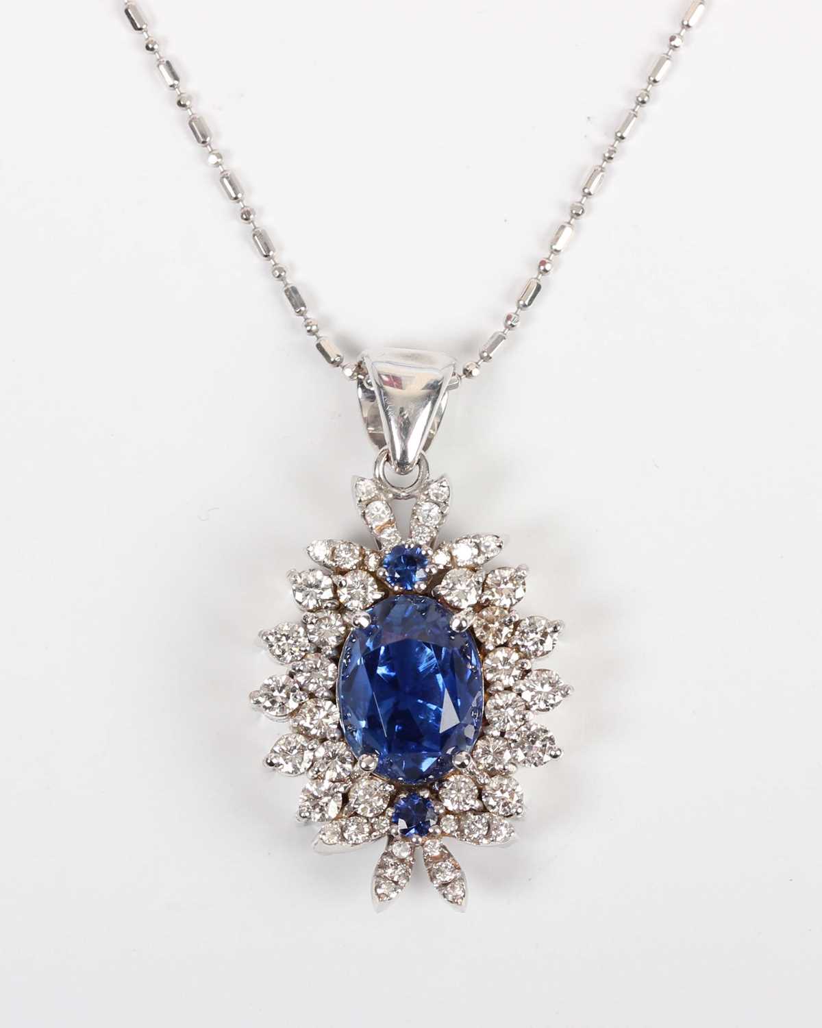 A white gold, sapphire and diamond cluster pendant, claw set with the principal oval cut sapphire